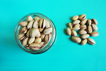 many grains of pistachios in a small glass jar next to the poured grains on a turquoise background top view