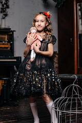 A little girl in a black dress with a red flower in her hair hugs a doll she took out of a cage.