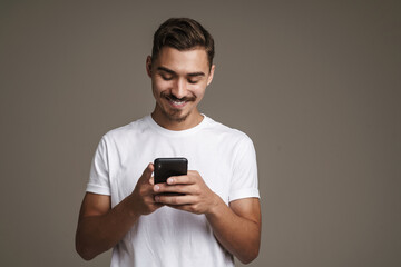Image of joyful unshaven guy smiling and using cellphone