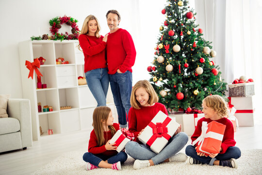 Photo of full big family five people meeting three little kids hold boxes ribbon bow guess present parents spectate near x-mas tree wear red jumper jeans in home living room indoors