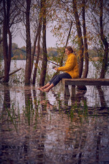 A barefoot Girl in a warm sweater sits with a fishing rod on a wooden bridge by the lake.