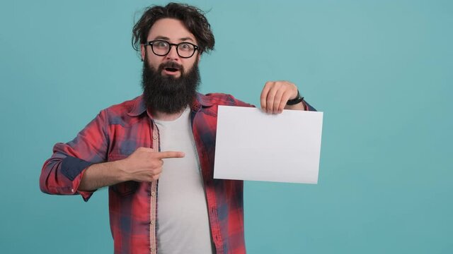 A bearded shocked young man points to a white sheet he holds in his hand, standing isolated over turquoise background.