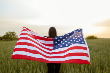 young happy beautiful  american  girl in pink top and jeans shorts with national USA flag  in green wheat field at sunse