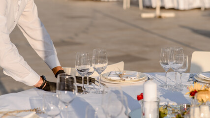 waiters hands in protective black gloves arrange a wedding party reception table decorated with flowers: plates, forks, knives and wine glasses.