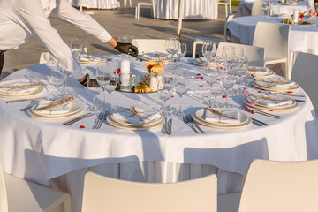 waiters hands in protective black gloves arrange a wedding party reception table decorated with flowers: plates, forks, knives and wine glasses.