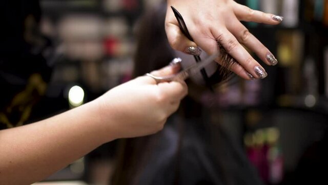 Close up of female hairdresser makes hairstyle on young brunette woman in salon cutting hair with scissors.