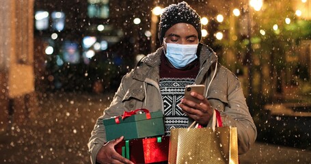 Portrait of man standing and texting on mobile phone. Young African American happy male with little Christmas tree and shopping bags typing on smartphone while snowing. Winter holidays concept