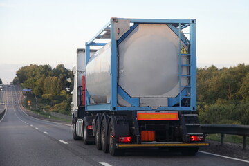 Gray three-axle semi truck with liquid acid barrel tank container frame moving on empty suburban two lane asphalt highway road on a summer day on green forest background, dangerous cargo logistics