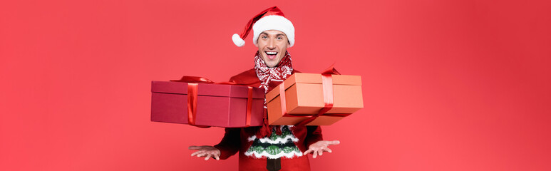 Cheerful man in santa hat throwing presents on red background, banner