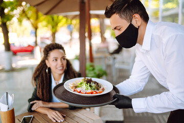Young waiter in protective face mask and gloves while bringing food to a customer in cafe during coronavirus outbreak. Waiter serving in motion on duty in restaurant.