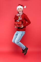 Cheerful man in santa hat holding gift box while jumping on red background
