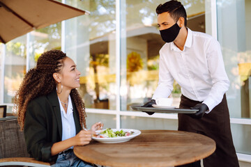 Young waiter in protective face mask and gloves while bringing food to a customer in cafe during coronavirus outbreak. Waiter serving in motion on duty in restaurant.