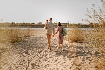 Happy family walking on sandy beach of river. Father, mother holding baby son on hands and going together. Rear view. Family Ties concept.