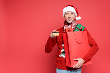 Positive man in santa hat holding shopping bag with presents and credit card on red background
