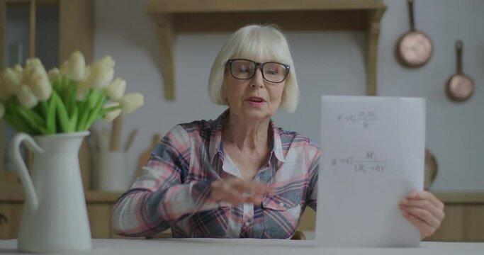 Senior teacher in glasses showing sheet of paper with physics equations looking at camera. Online education and distance learning process. Webcam view of working from home woman talking. - Powered by Adobe