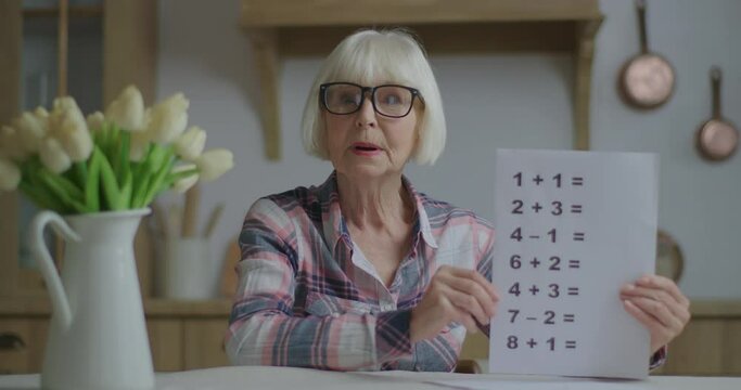 Senior teacher in glasses showing sheet of paper with simple mathematical equations looking at camera. Online education and distance learning process. Webcam view of working from home woman talking. - Powered by Adobe