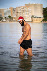 Fototapeta na wymiar Tall fit guy stands at the middle of the water in Palmanova beach (Mallorca, Spain) while smiling and wearing a Santa Claus hat during winter time