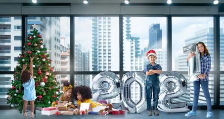 Children of different color and races have fun with metallic silver number balloons and Christmas trees, 2021 Happy new year,  against building background