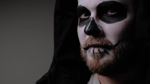 Man, disguised as a zombie, with a beard and a black hood on his head, raises his head and stares into the camera with a seriously aggressive look. Halloween eve. October holiday.