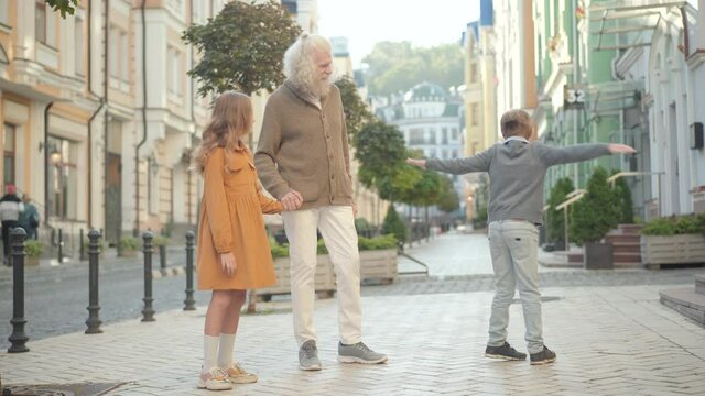 Wide shot of happy Caucasian grandfather strolling with cute boy and girl outdoors on sunny summer day. Cheerful grey-haired senior man and positive grandchildren walking along street in city.