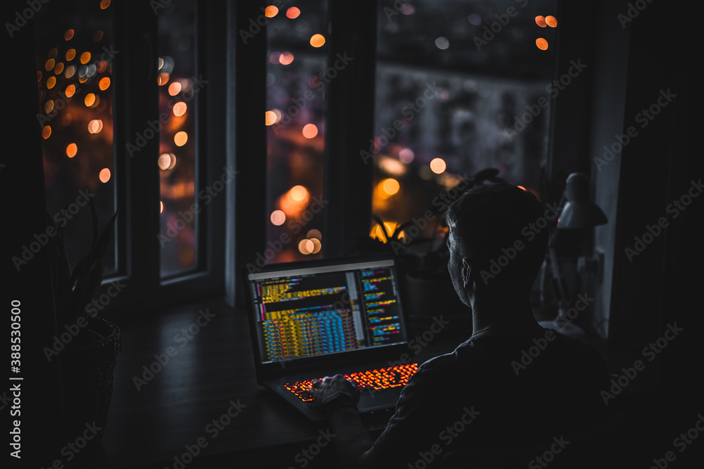 Wall mural a young man programmer coding on a laptop in the dark with a view of the lights of the night city, c