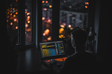 A young man programmer coding on a laptop in the dark with a view of the lights of the night city, color lighting in the room, home decor