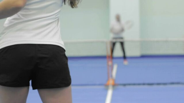 A young woman in a white t-shirt and black shorts plays tennis in the gym. The view from the back.