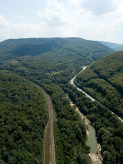 Aerial view of green river between railway and highway road with cars, surrounded by mountains, forest, green trees, sky, clouds. Caucasus nature landscape, Krasnodar Krai, Russia.