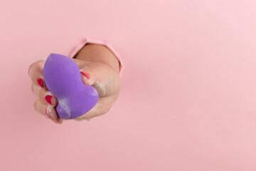 Girl hand holds a beauty blender for makeup from a hole in a pink background, copy space. Sponge in soapy foam, how to clean.