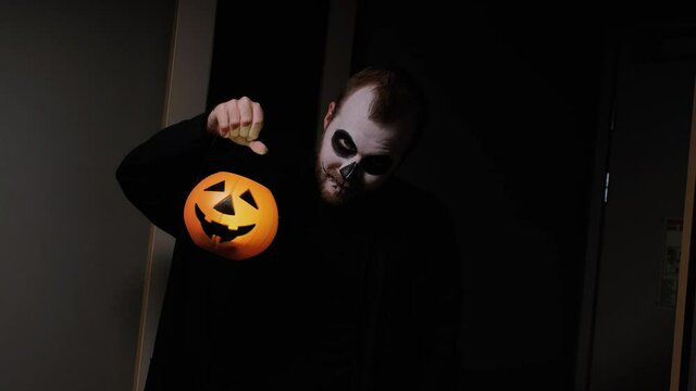 A man disguised as a zombie, with a beard and in black clothes, looks seriously into the camera while holding a lantern in the form of an orange pumpkin head. Halloween Eve. October holiday.