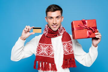 Smiling man wearing scarf and sweater, showing wrapped present and credit card isolated on blue