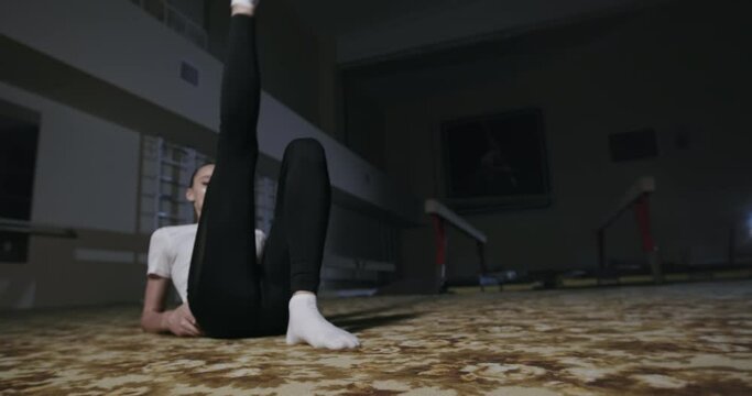 Female gymnast stretching on floor. Slim young woman lying on floor and lifting leg while doing stretching exercise during gymnastic training in dark gym