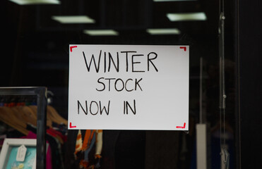 Winter Stock NOW IN sign hand written on white A4 paper suspended in the window of a local business. Advising people to still come inside even with the illness and pandemic coming