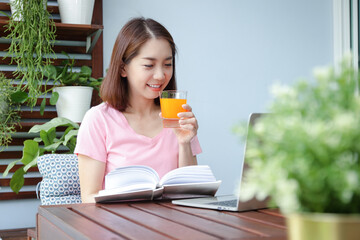 A happy smiling asian woman holding a glass of orange juice And read books Sit back and relax on the balcony. Concept of health problems for women in their 40s. Eat a good and healthy diet.