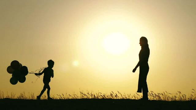 Silhouette of a happy family. A child with balloons runs to his mother. Mother hugs her son.