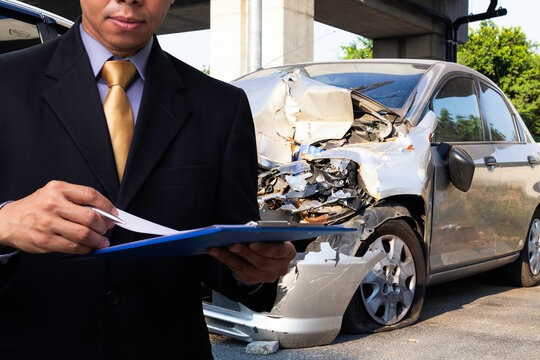 Side view of insurance officer writing on clipboard while insurance agent examining silver car after accident.
