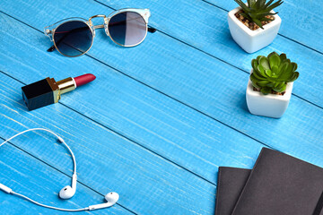 Sunglasses, white earphones, red lipstick, two black folders and green succulents in pots on blue wooden desktop or background. Close up, copy space