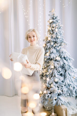 Obraz na płótnie Canvas Christmas, winter holidays concept. Beautiful charming blond woman in trendy clothes, posing in luxurious decorated Christmas studio with Christmas tree, holding white present box