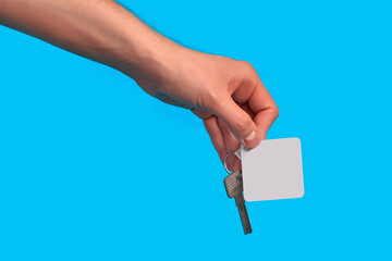 Hand of unknown male is holding a key with blank white square plastic key fob on metal ring against blue studio background. Close up, copy space