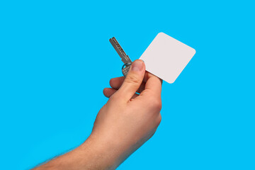 Hand of unrecognizable guy is holding a key with empty white square plastic key fob on metal ring against blue studio background. Close up, copy space