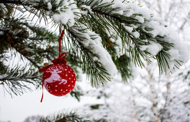 Christmas background with a red knitted Christmas ball with deer on snow-covered pine branch in winter forest with copy space. New year holiday, Christmas celebration, greeting card. Merry Christmas