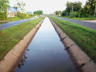 A canal with a road on both sides. The background has green trees on the sides and white sky. Selective focus