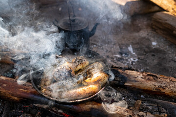 Large chunks of freshly caught fish are fried in a skillet over a campfire, next to a kettle, in a camping, on a summer day. 