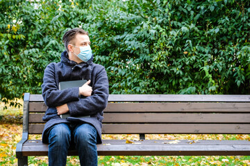 Handsome young European man in a park with laptop with a medical face mask on. Freelance working outside the office during an Covid-19 epidemic. Selective focus