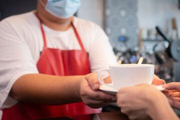 Selective focus of coffee cup and background of Asian barista woman wearing face masks to prevent contagious diseases and serve to customers in the coffee shop. The concept of prevention from COVID 19