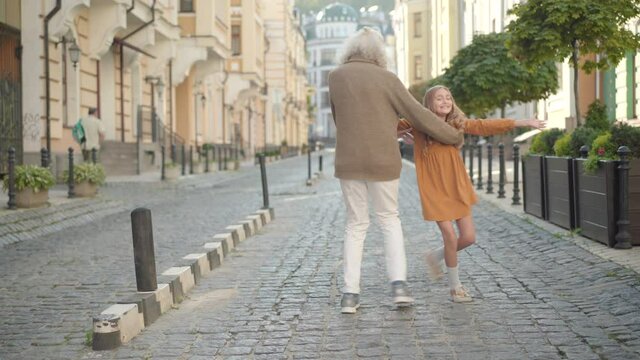 Wide shot portrait of cheerful senior man and cute little girl jumping and dancing on city street. Happy carefree grandfather and granddaughter having fun on sunny day outdoors and leaving.