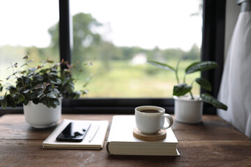 White coffee mug and phone with notebooks and plant pot on brown wooden table in front of window
