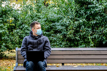 Handsome young European man in a park with laptop with a medical face mask on. Freelance working outside the office during an Covid-19 epidemic. Selective focus