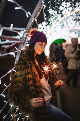 The beautiful young lady standing near Christmas fair with sparkles in her hands 