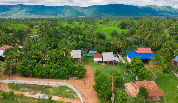 A Top Down Aerial View Of A Small Country Town With Traditional Houses With Orange Roofs, A Red Dirt Road, Rice Fields, And Palm Trees In The Jungle In Cambodia.